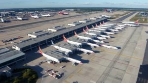 Aerial photograph of Hartsfield-Jackson Atlanta International Airport showing terminal buildings, multiple aircraft parked at gates, and ground service vehicles on the tarmac during daytime with clear skies