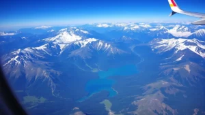 Aerial view of snow-capped Rocky Mountain peaks with emerald green forests and pristine lakes visible below, taken from airplane window during daylight