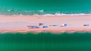 Aerial view of turquoise waters and white sand beach at Destin, Florida coastline with colorful beach umbrellas and tourists enjoying the shore during golden hour sunset