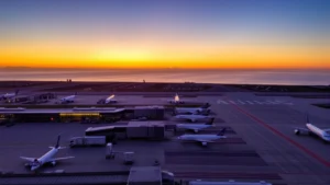 Aerial view of LAX terminal at sunset with planes lined up at gates, Pacific Ocean visible in background, realistic photography, no text visible