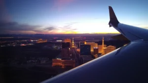 Aerial view of Las Vegas Strip at sunset with desert landscape, modern aircraft wing visible in frame, golden hour lighting, vibrant city lights beginning to twinkle below