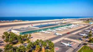 Aerial photograph of Blaise Diagne International Airport in Dakar, Senegal with modern terminal buildings and aircraft parked at gates, surrounded by palm trees and coastal landscape under blue sky